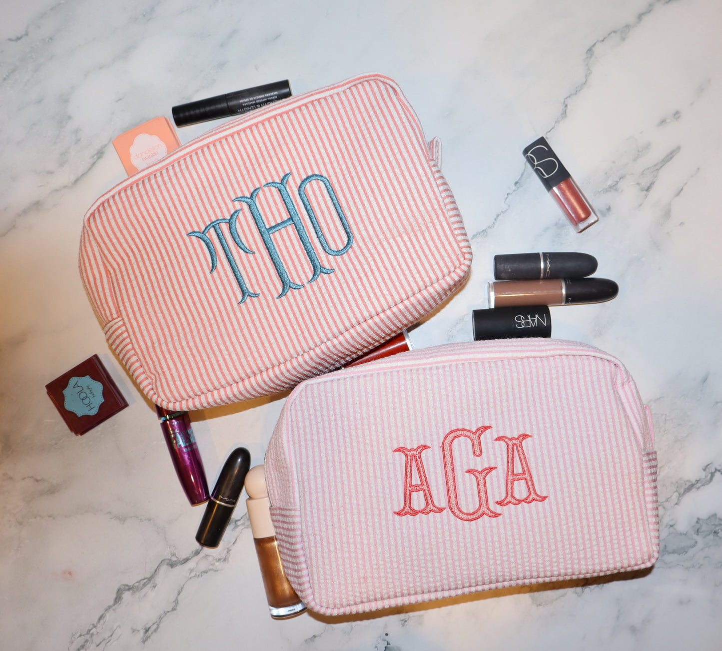 Two pink cosmetic bags with monogrammed initials on a marble surface with makeup items around.