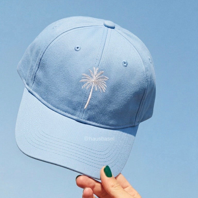 A light blue baseball cap with a white embroidered palm tree, held by a hand against a clear blue sky.