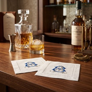 Two white linen coasters with blue embroidered monograms resting on a wooden bar counter, surrounded by whiskey bottles, a decanter, and a glass of amber liquor in a bar setting.