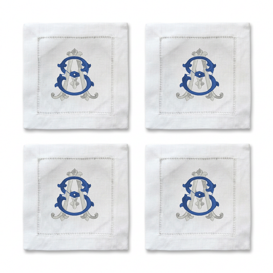 Four white embroidered cocktail napkins arranged in a 2x2 grid, each featuring a blue and silver monogram crest with a white veil motif.