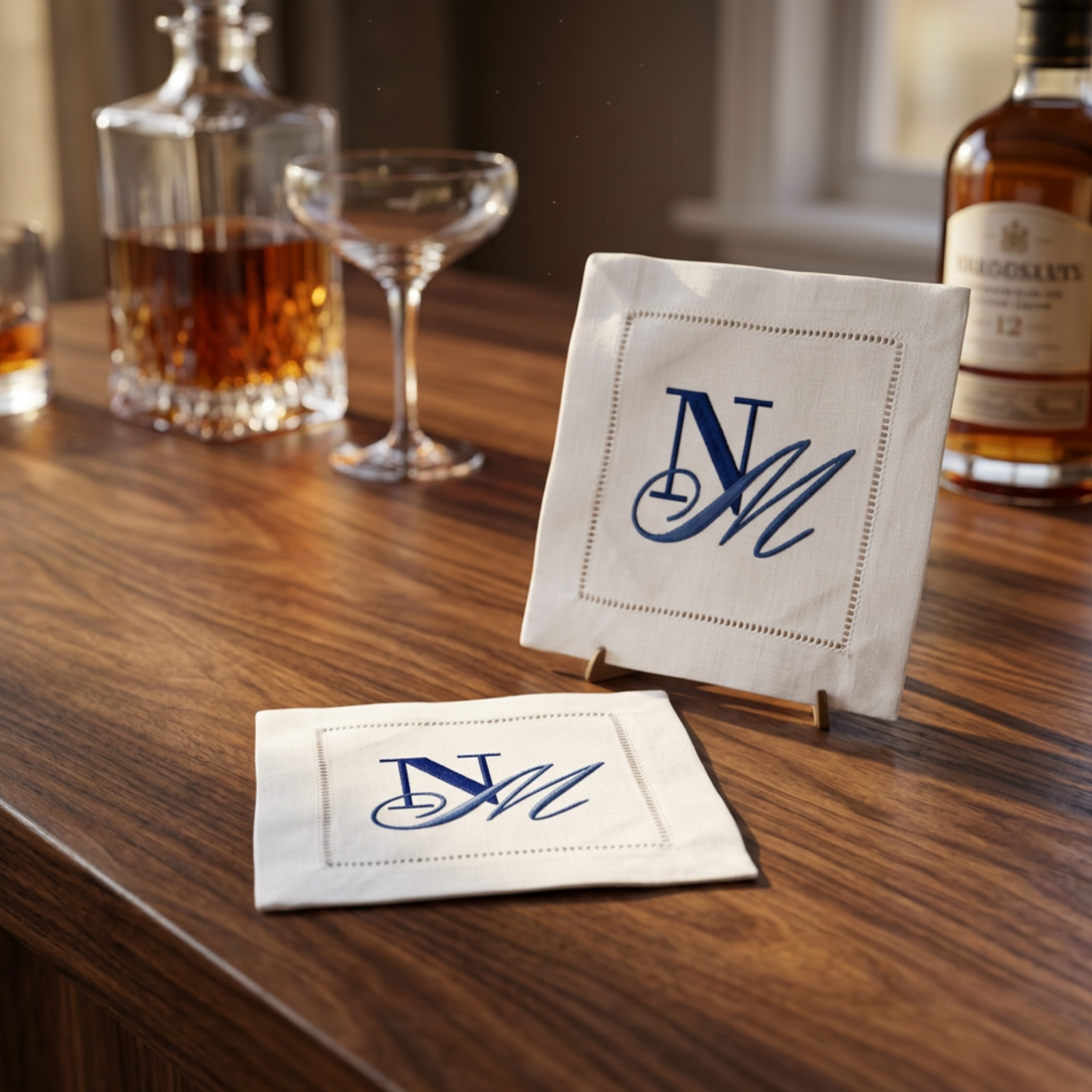 Two white embroidered monogram coasters with blue 'N M' script, displayed on a wooden bar table with a decanter, coupe glass, and whiskey bottle in the background.