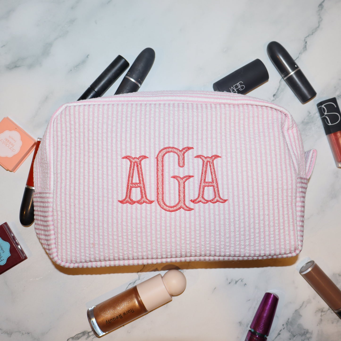 Pink corduroy pouch with 'AGA' monogram surrounded by makeup items on a marble surface