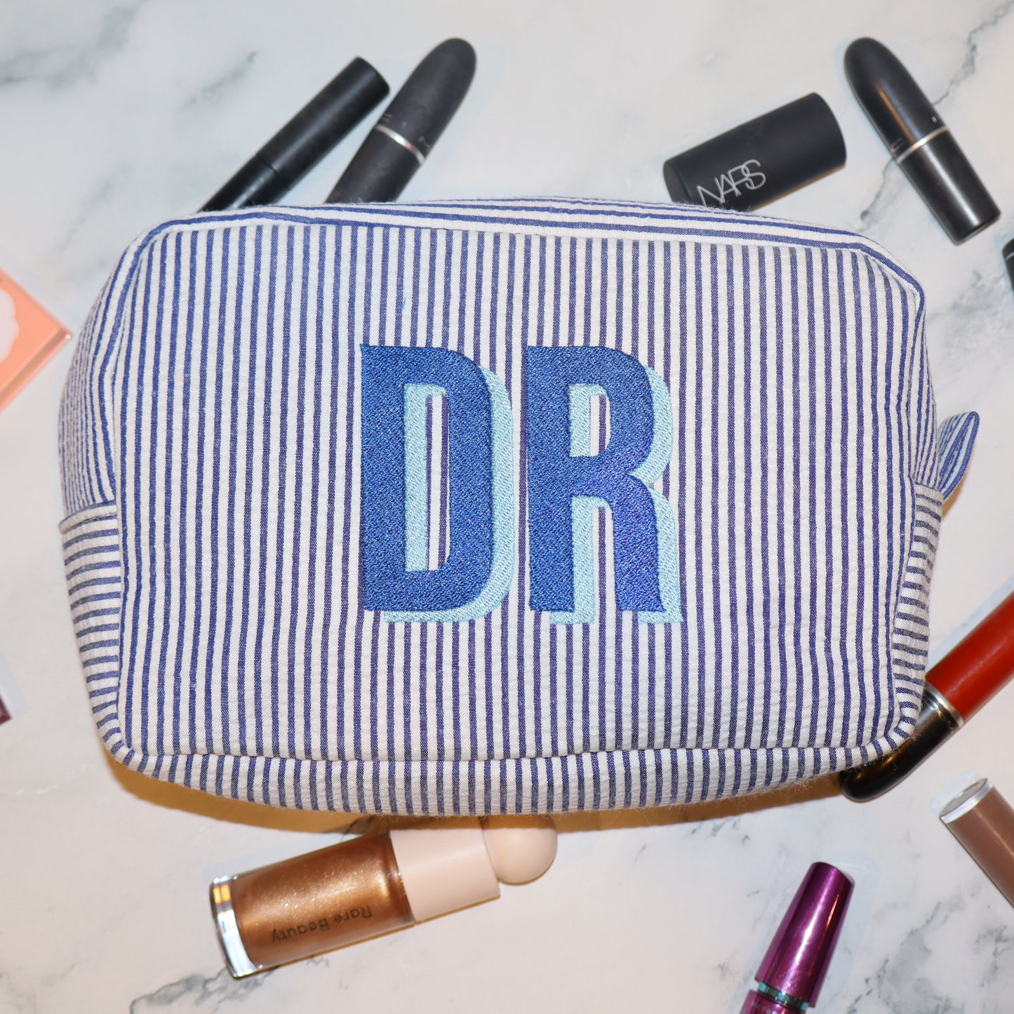 Makeup bag with 'DR' initials surrounded by makeup items on a marble surface