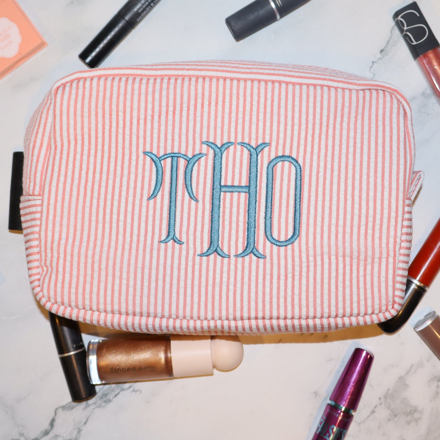 Pink and white striped makeup bag with 'THO' monogram surrounded by makeup items on a marble surface.