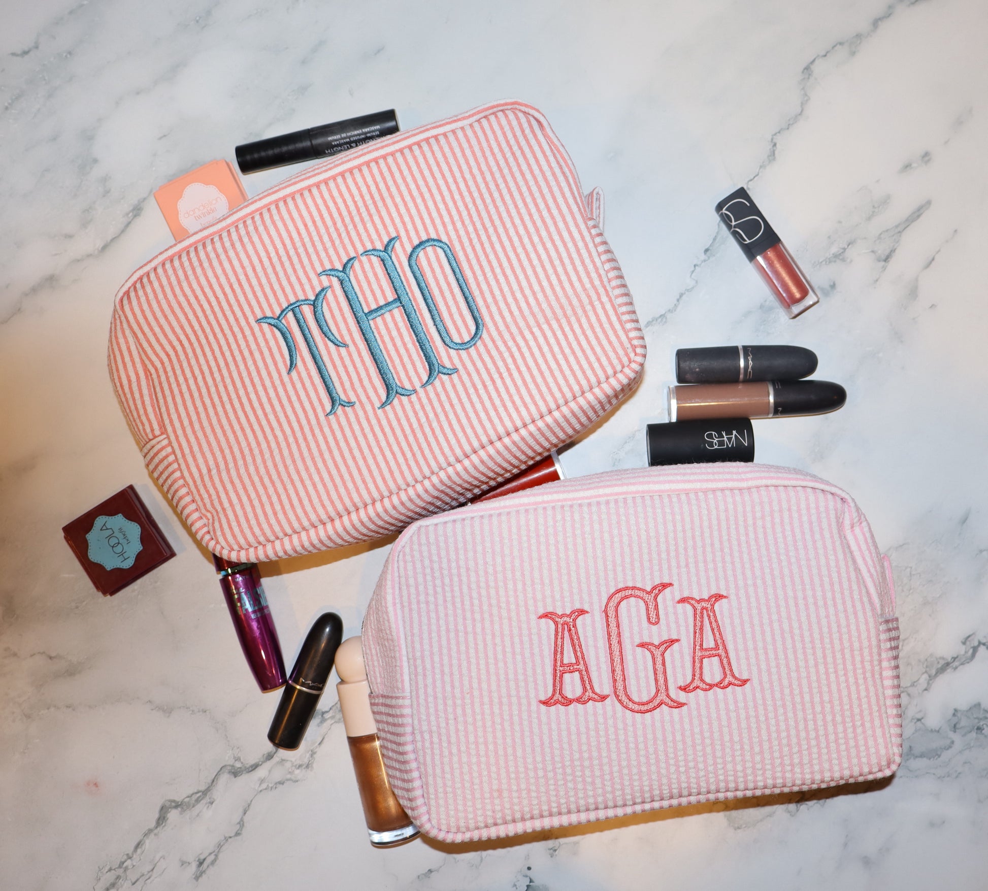 Two pink cosmetic bags with monogrammed initials on a marble surface with makeup items around.