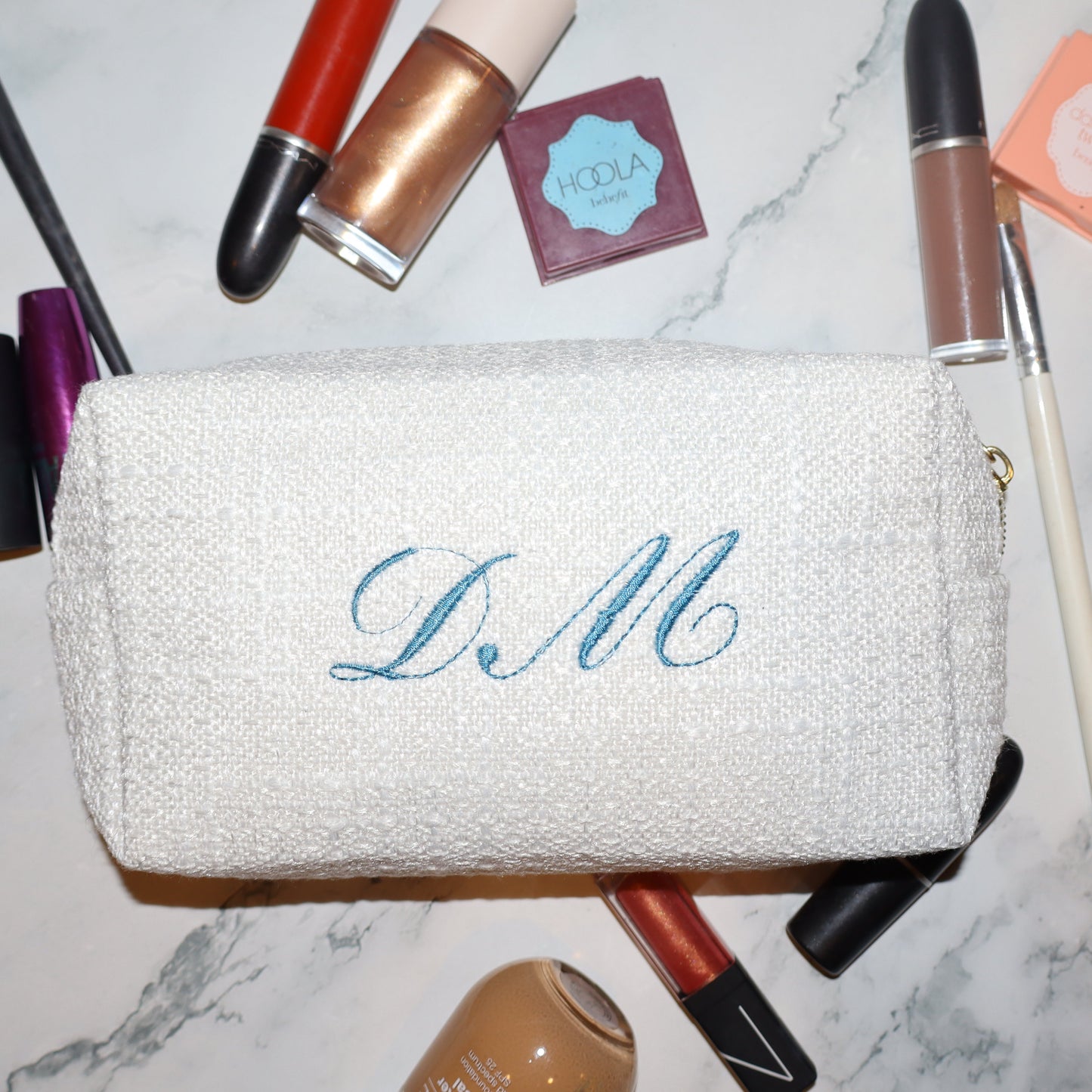 White makeup pouch with 'DM' monogram surrounded by makeup items on a marble surface