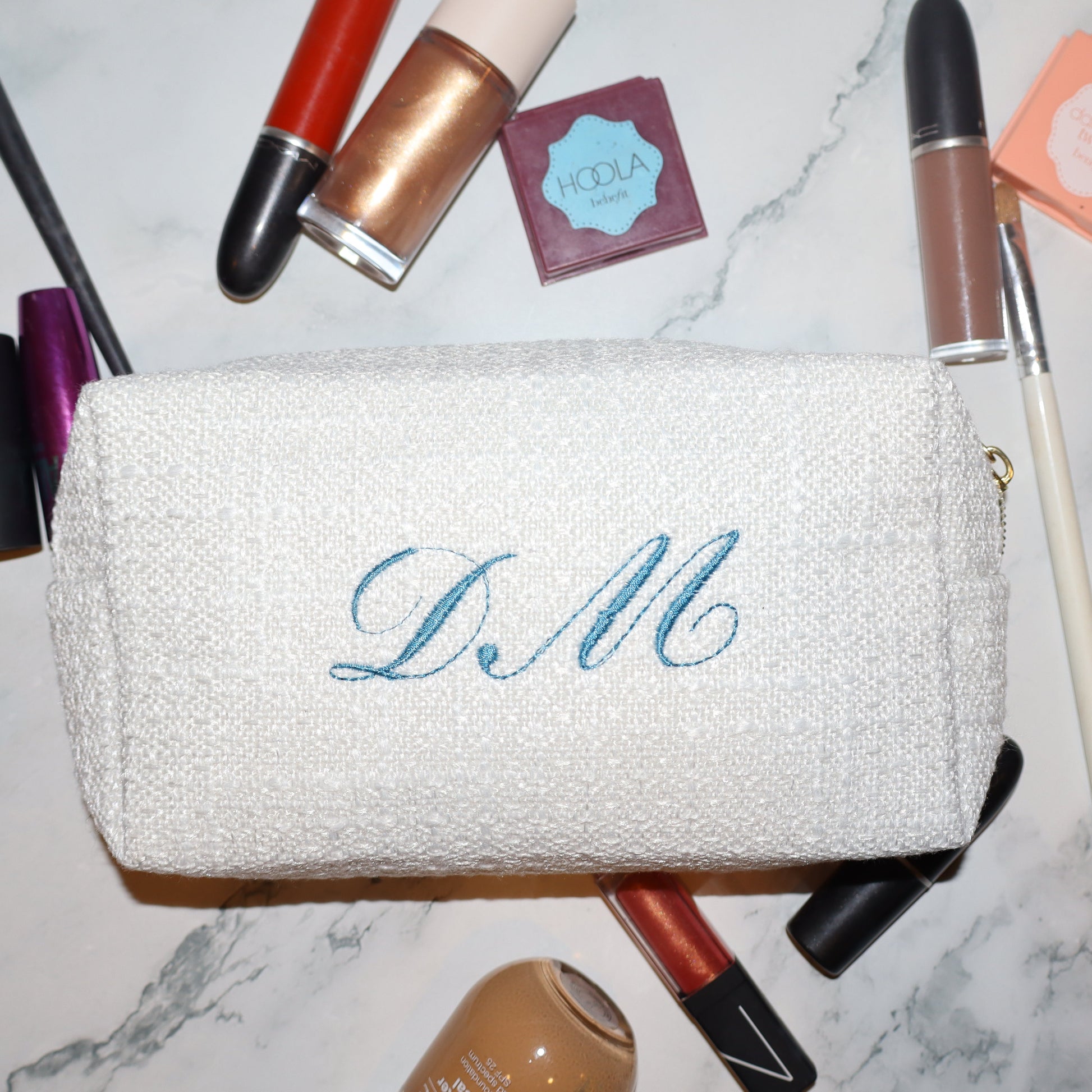 White makeup pouch with 'DM' monogram surrounded by makeup items on a marble surface