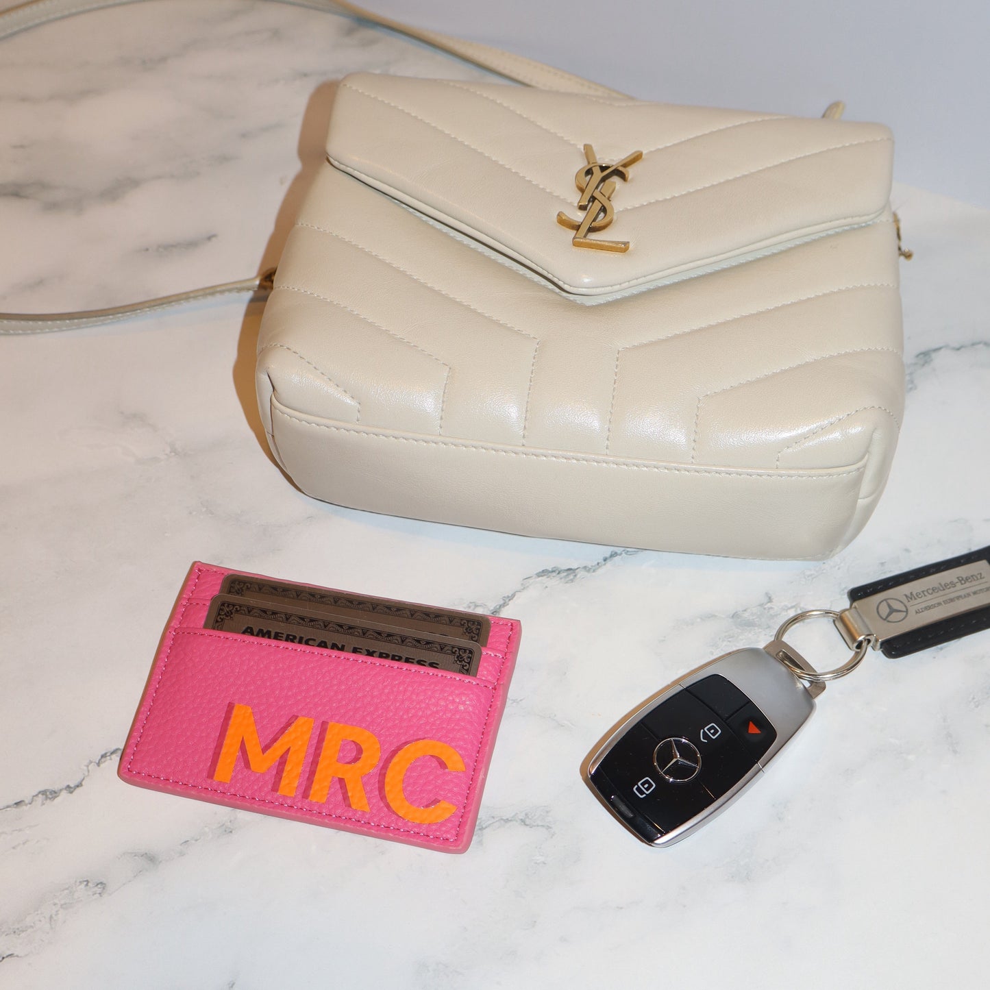 Beige handbag, pink cardholder with 'MRC' initials, and Mercedes-Benz key fob on a marble surface