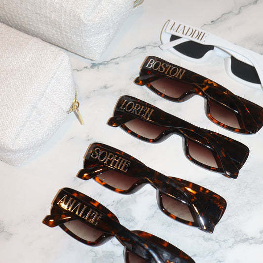 Set of tortoiseshell sunglasses with personalized names on a white marble surface
