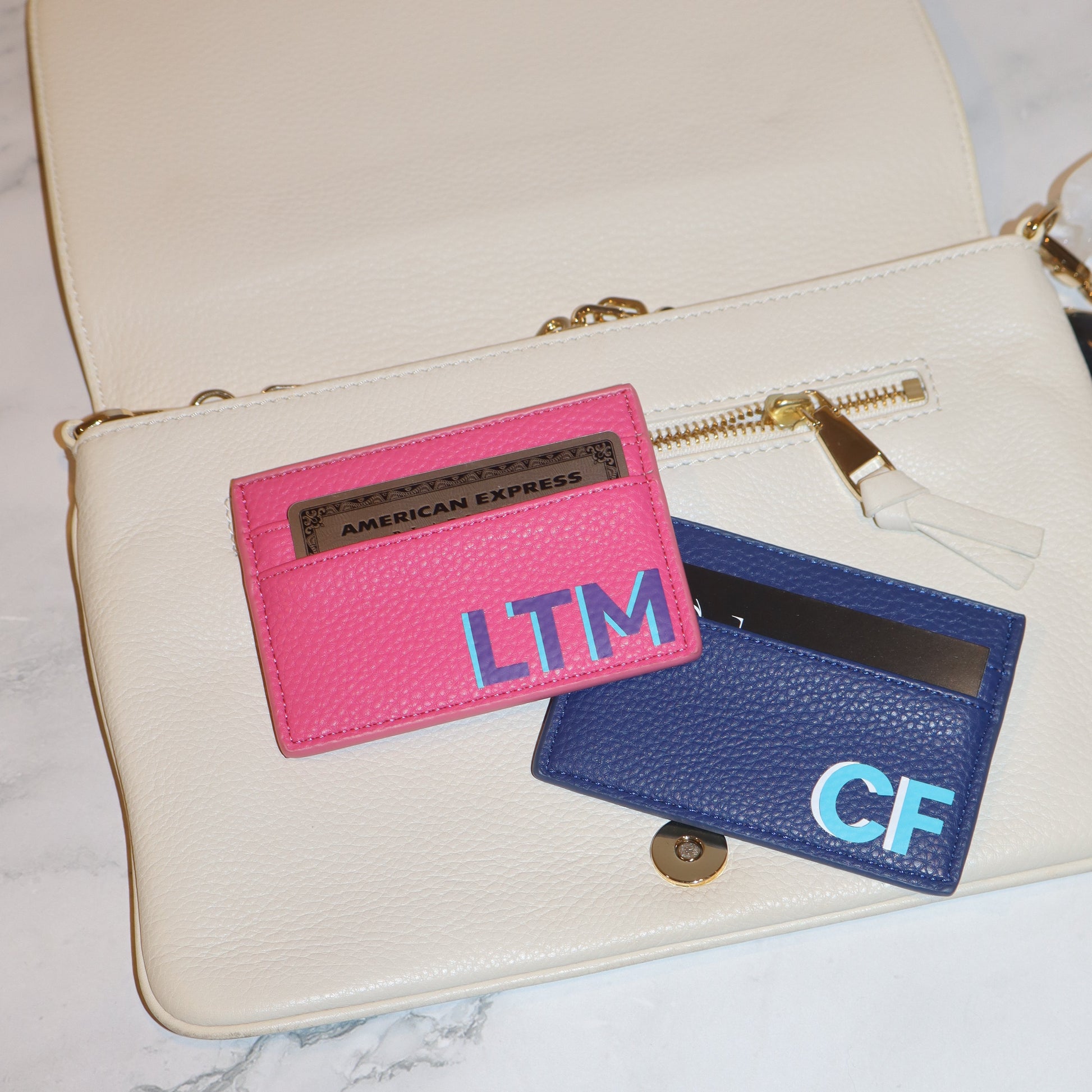 Twowallets, one pink and one blue, with initials on a marble surface.