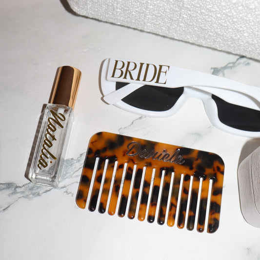 Tortoiseshell comb, bottle of perfume, and white sunglasses on a marble surface.