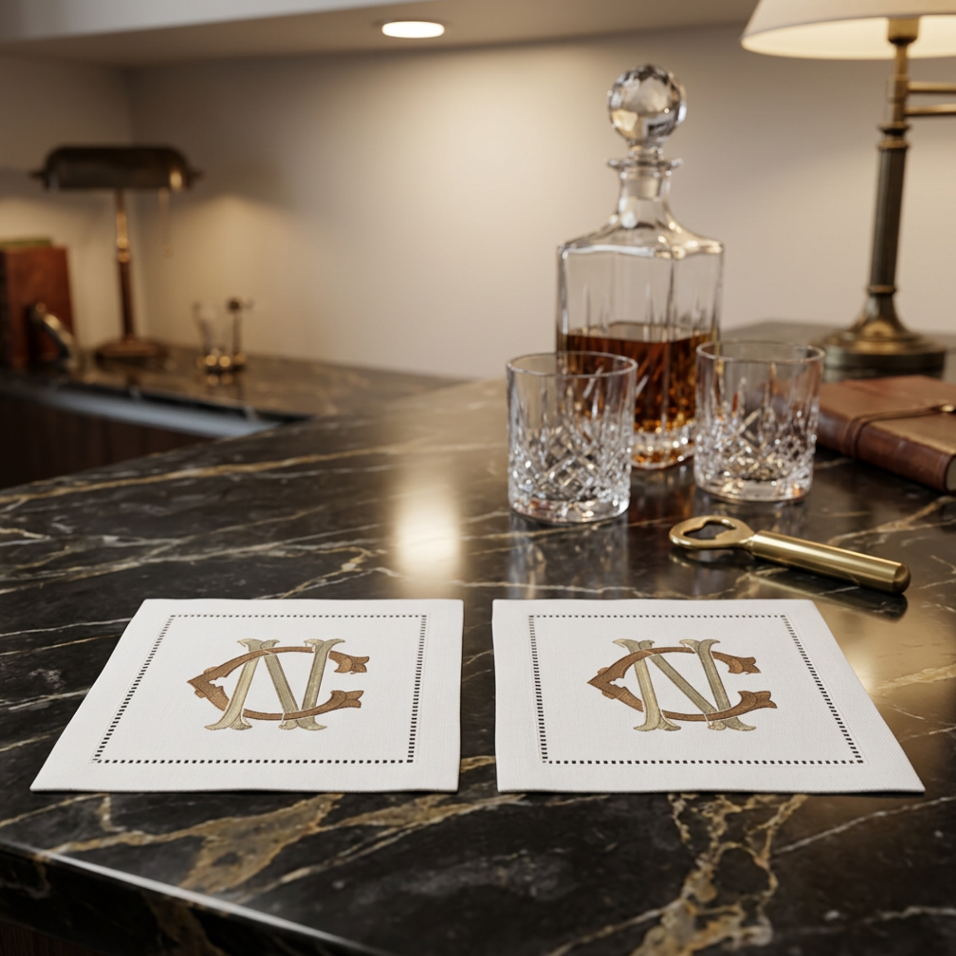 Two white square napkins with gold monogrammed lettering and dotted borders are arranged on a dark marble bar counter next to a glass decanter, two cut-glass tumblers, and a brass bottle opener in a dimly lit indoor setting.
