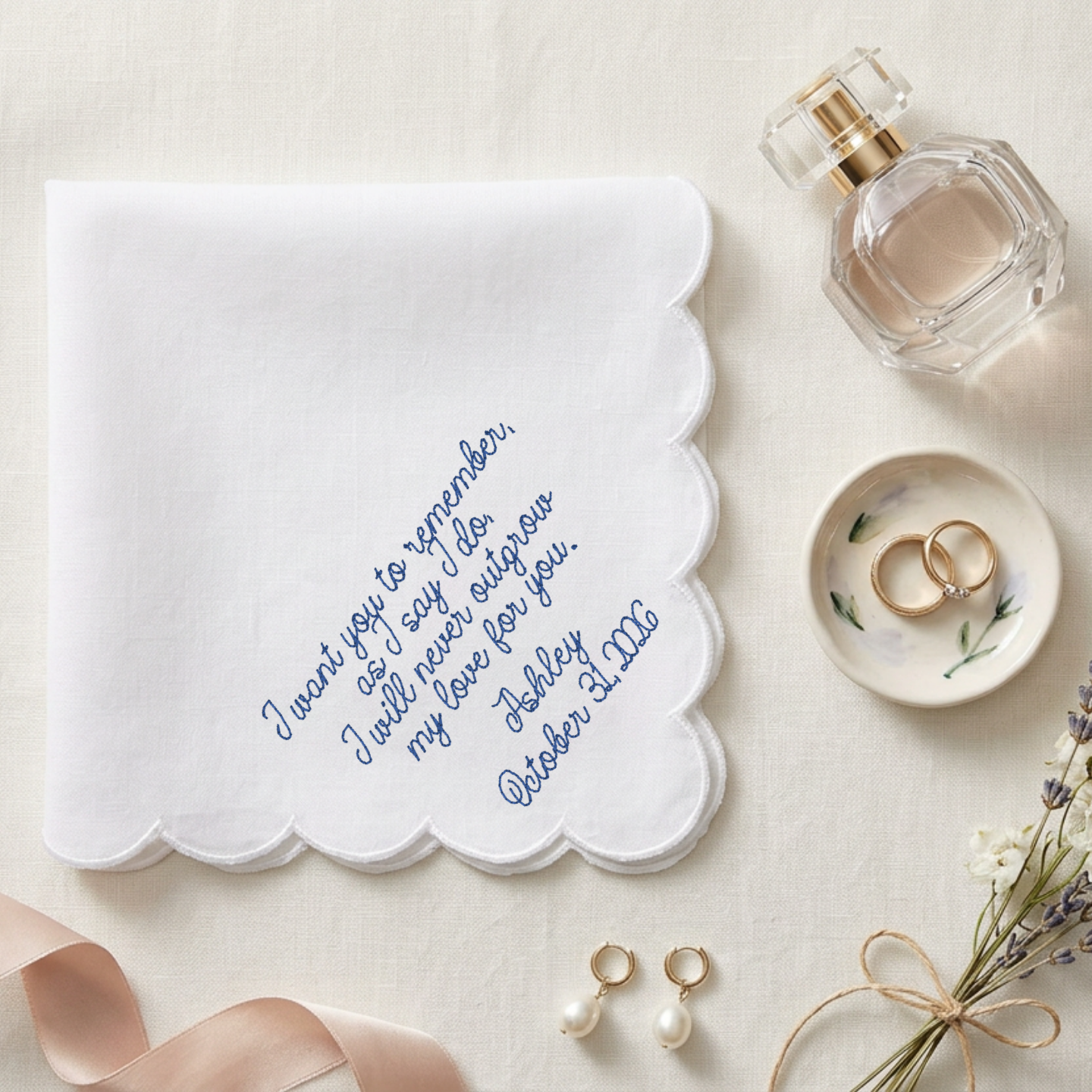 A white embroidered handkerchief with scalloped edges lies on a beige fabric surface, styled with a perfume bottle, rings in a dish, pearl earrings, a ribbon, and dried flowers. The handkerchief has blue embroidered text reading, 'I want you to remember, as I say this, I will never forget our my love for you, Ashley October 21st.